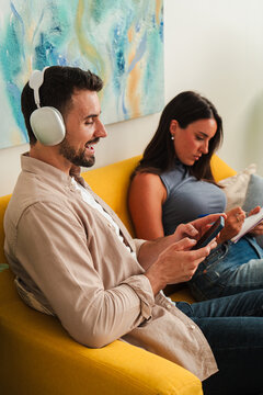 A relaxed couple sits on a yellow couch using a tablet and headphones, enjoying a private digital break. The composition captures calm mood, focused interaction, and comfortable home lifestyle.
