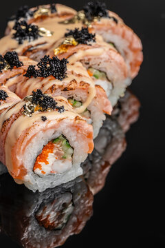 Close view of assorted sushi rolls with salmon, rice, vegetables, creamy sauce, black roe, and edible gold arranged on a reflective dark surface, Japanese cuisine