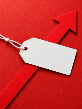 Blank white price tag tied with string lies over a red upward arrow on a textured red background, suggesting price growth, promotion, or sales success.