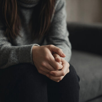 Young woman in a gray sweater sits on a sofa with her hands clasped, conveying worry, stress, or quiet reflection in an indoor setting.
