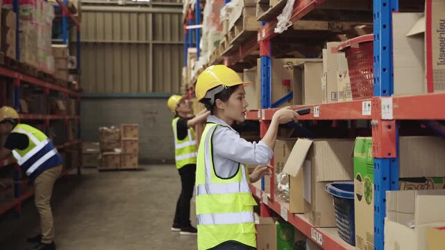 Team of Asian warehouse workers in uniform with yellow safety helmets holding and using a barcode scanner for checking inventory stock on shelves together in a logistics distribution factory center. 