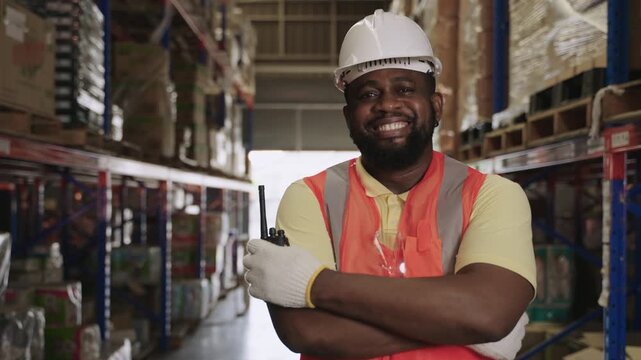 Portrait of a happy African American male warehouse worker in an orange vest and white safety helmet holding a walkie-talkie standing with arms crossed while looking at camera in distribution factory.