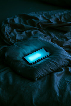 Minimalist glowing blue smartphone on dark bed with moody low key lighting cold cyan