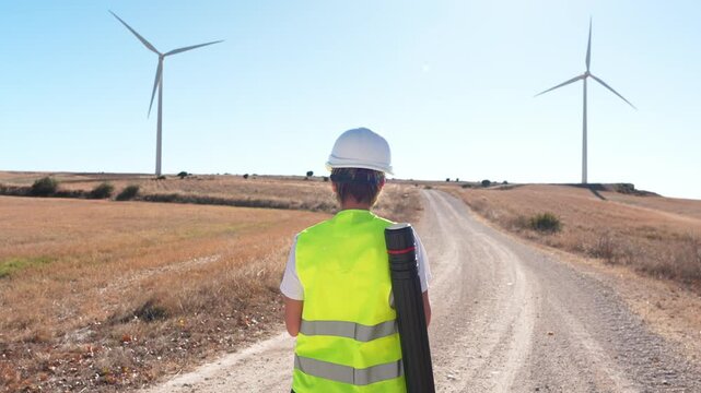 Female engineer in safety vest and hard hat walking on a rural road towards a wind farm. Sustainable energy and professional work concept