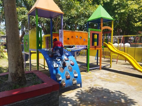 A young girl plays on a colorful public playground in Java, climbing a small rock wall surrounded by trees and outdoor recreational equipment.