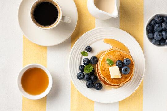 Golden fluffy pancakes topped with butter, blueberries and fresh mint on a white plate, accompanied by coffee and syrup on a light stone background.