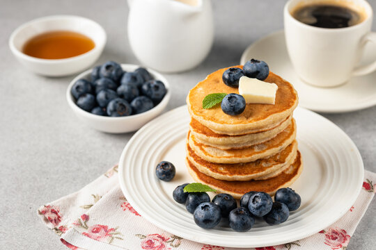 Golden fluffy pancakes topped with butter, blueberries and fresh mint on a white plate, accompanied by coffee and syrup on a light stone background.