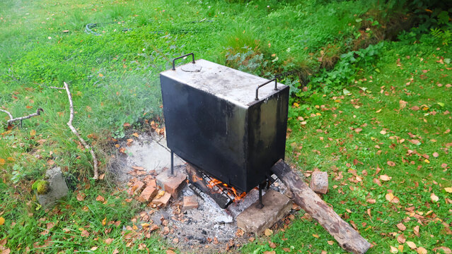 Rustic metal smoker billowing smoke over a small wood fire in a grassy backyard strewn with fall leaves, cooking meat outdoors for a traditional autumn cookout