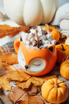 Creamy pumpkin spice latte topped with marshmallows and spices in Cute orange fox mug, surrounded by pumpkins and colorful autumn leaves.