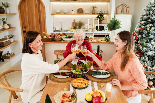 Three people toast at a festive holiday dinner in a cozy home, with a Christmas tree, roast dishes, and desserts, celebrating togetherness.
