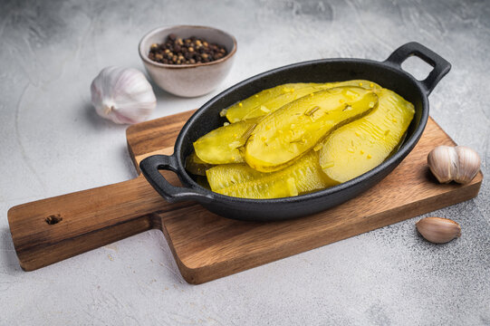 Rustic pickled cucumber slices in cast iron dish with garlic and spices on textured light background, vibrant brined yellow food.