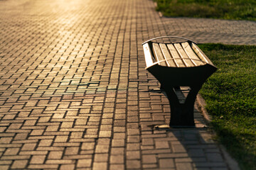 A lonely wooden bench stands on a paved stone walkway in a park during a warm and golden sunset.  © Dina Luk