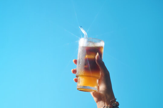 A hand holding a glass of iced coffee against a bright blue sky