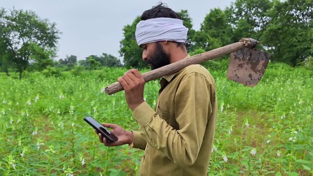 Indian farmer using mobile phone while carrying a hoe on his shoulder