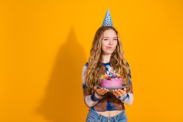 Young girl wearing a party hat holds a colorful cake against a bright yellow background