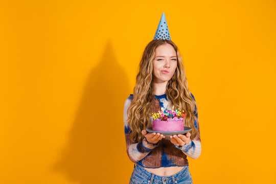 Young girl wearing a party hat holds a colorful cake against a bright yellow background