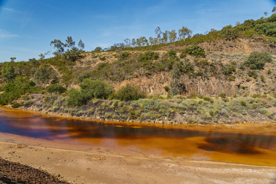 View of the orange-colored water of the river at Minas de Sao Domingos, surrounded by rocky terrain and sparse vegetation in Corte do Pinto, Beja District, Portugal.