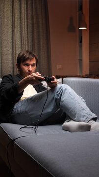 Vertical video young man focused on gaming, holding a controller while sitting comfortably on a modern sofa in his living room, enjoying entertainment and leisure time
