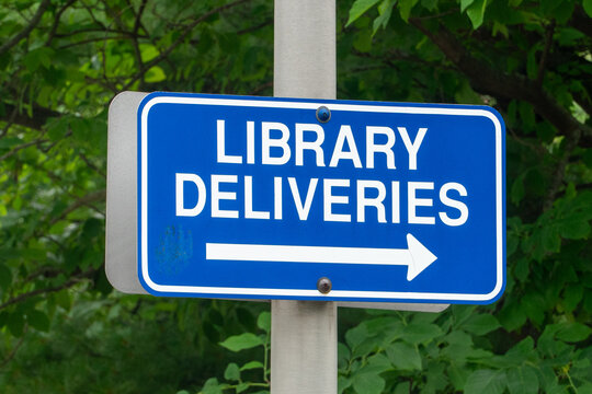 Blue Library Deliveries sign with a white arrow pointing right, mounted on a metal pole, indicating direction for efficient book and media transport logistics amidst lush green foliage.