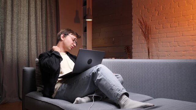 Young man yawning while working on a laptop at home, feeling tired and exhausted from remote work and overtime, struggling with sleep deprivation and burnout