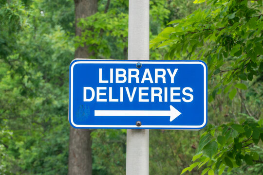 Blue 'LIBRARY DELIVERIES' sign with a white arrow pointing right, mounted on a pole amidst blurred green trees and foliage, indicating a specific service area or direction at an institution.