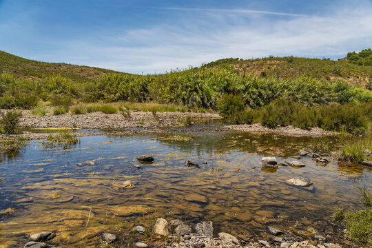 View of a shallow rocky stream flowing through the rural landscape with low hills and Mediterranean shrubs under a clear blue sky in Vaqueiros, Santarem, Portugal.