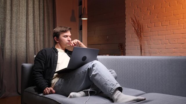 Young man sitting on a sofa with a laptop, holding his glasses and rubbing his tired eyes, feeling the symptoms of eyestrain and headache from prolonged screen time during remote work