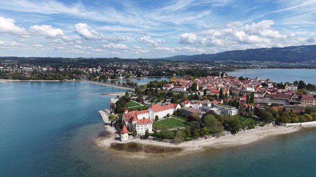 Aerial drone view of Lindau Island on Lake Constance in Bavaria, Germany, showing historic waterfront buildings, Mangturm tower, red tiled roofs and the old town surrounded by blue water.