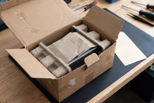High-angle view of an open corrugated cardboard box on a wooden workbench, revealing a new electronic device cushioned by recycled molded pulp and kraft paper, suggesting unboxing.