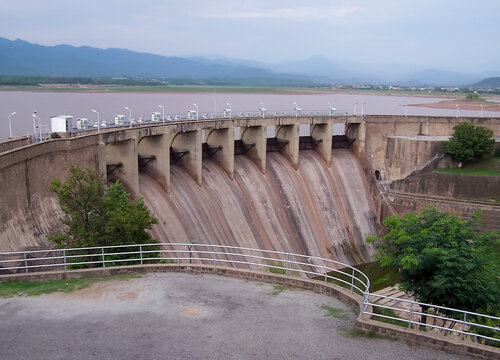 View of Rawal Lake dam with its concrete spillway structure, calm water, and distant mountains under an overcast sky in Islamabad, Islamabad Capital Ter, Pakistan.