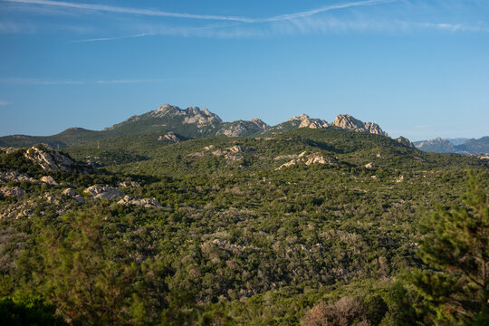 View of the rugged mountainous landscape characterized by rocky peaks, dense Mediterranean scrub, and green hills under a clear blue sky in Marinella, Sardegna, Italy.