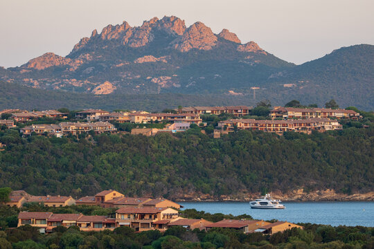View of the coastal resort with Mediterranean architecture, lush green hills, rocky mountains in the background, and a luxury yacht anchored in the calm blue bay in Marinella, Sardegna, Italy.