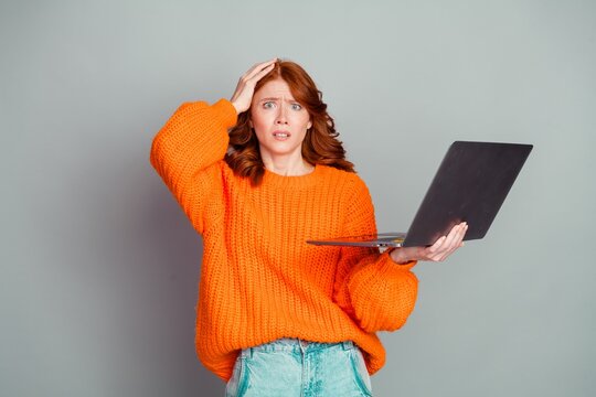 Confused young redhead woman in orange sweater holding laptop and scratching her head, frustrated by a computer error or unexpected online problem