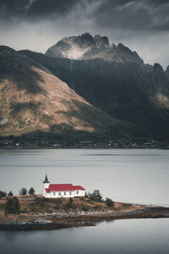 View of Sildpollnes kirke with its red roof situated on a small peninsula by the water, framed by dramatic mountains under a moody, overcast sky in Laupstad, Nordland, Norway.