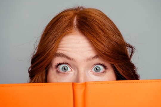 Surprised redhead woman peeking over orange notebook with wide eyes and playful expression