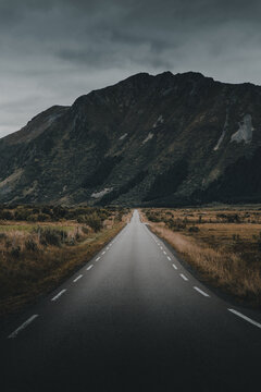 View of a long, straight asphalt road leading towards a dramatic, dark mountain under an overcast sky in the rugged landscape of Lofoten, Nordland, Norway.
