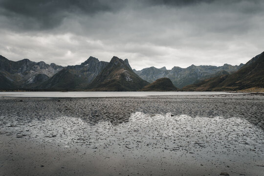 View of the rugged mountains along Midnattsolveien reflected in the tidal flats under a dramatic cloudy sky in Laukvik, Nordland, Norway.