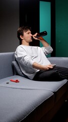 Fototapeta premium Young man lounging on a couch at home, holding a beer bottle and tv remote, relaxing in his living room and unwinding during an evening or weekend downtime