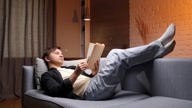 Young man relaxing on a modern sofa at home, feet up and reading a book in warm evening light, cozy living room setting conveying calm, leisure and contentment