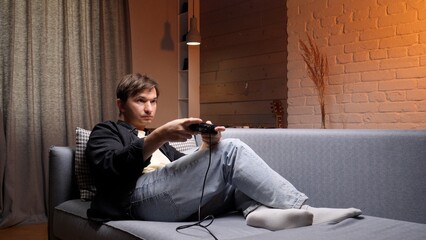 Young man engrossed in playing a video game using a wired controller, sitting comfortably on a gray sofa in a dimly lit living room during the evening, enjoying his leisure time © Aliaksandr