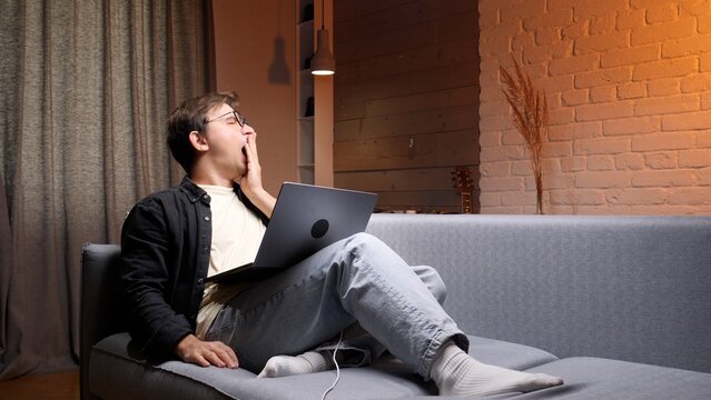Young man on a sofa yawning while working late on a laptop at home, showing exhaustion and burnout from long hours, sleep deprivation and remote work fatigue