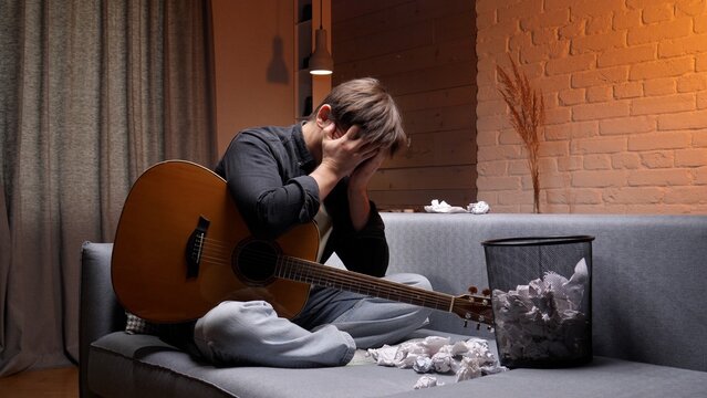 Man sits on a sofa, covering his face with his hands, feeling stressed and frustrated with a creative block while an acoustic guitar rests beside him and crumpled papers fill a waste basket