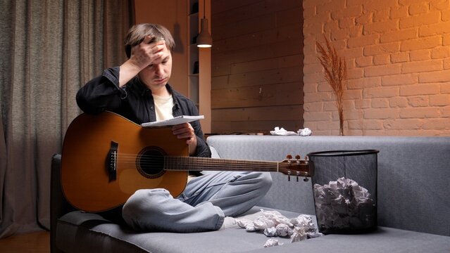 Young man on a sofa holding an acoustic guitar and crumpled paper, frustrated and pensive during a bout of writer's block while trying to compose new songs