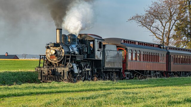 View of a vintage steam locomotive pulling passenger cars through a green field with smoke billowing from the stack in Ronks, Pennsylvania, United States.