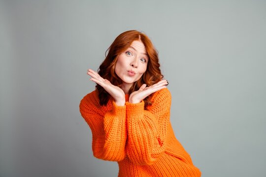 Playful redhead woman in orange sweater posing with a puckered kiss and hands framing her face studio portrait showing cheerful, quirky expression and friendly mood