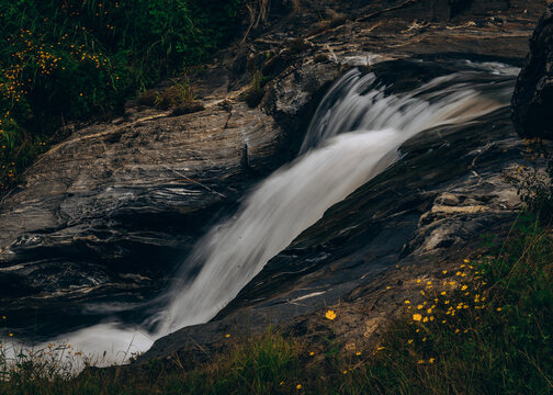 View of a cascading waterfall flowing over dark, textured rock formations surrounded by lush green vegetation and small yellow wildflowers in Jos East, Plateau, Nigeria.