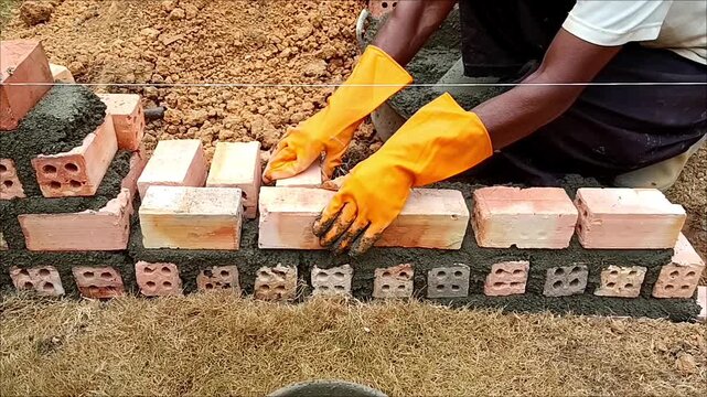 Construction worker laying red bricks and applying fresh mortar during the foundation construction of a house, highlighting skilled masonry work and the early stages of residential construction