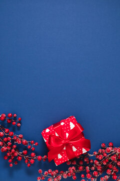Vertical Christmas background with red gift box and frosty berry branches on blue backdrop. Festive winter holiday bottom border composition with large copy space for text, top view flat lay.