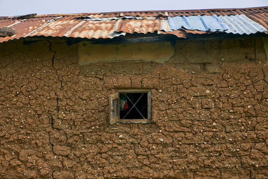 View of a rustic mud-brick wall with a small open wooden window and a weathered corrugated metal roof in Radung, Plateau, Nigeria.