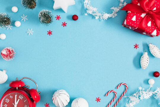 Christmas and New Year frame background with red vintage alarm clock, gift box, mini trees, and snowflakes on light blue backdrop. Festive winter holiday border flat lay with central copy space.